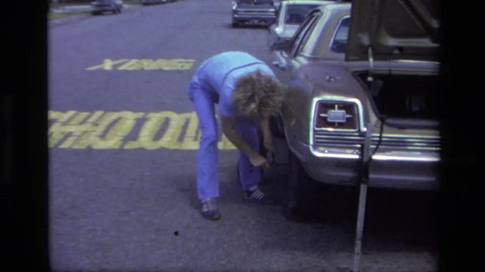 In 1979 Laguna Beach home-movie footage, a woman in blue bends near the rear wheel of a station wagon with the trunk open on a street.