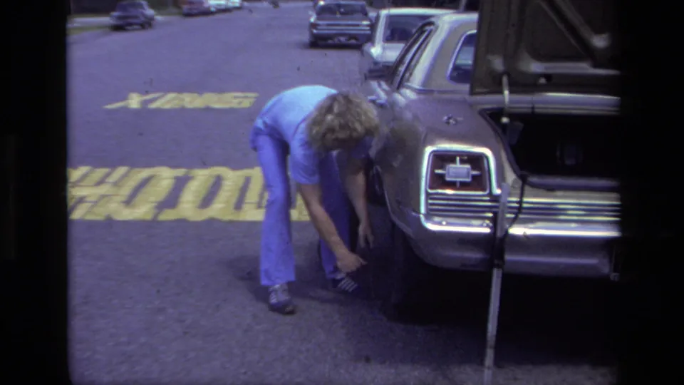 The woman remains bent over the station wagon's rear wheel, with the open trunk, car emblem area, and painted street lettering visible.