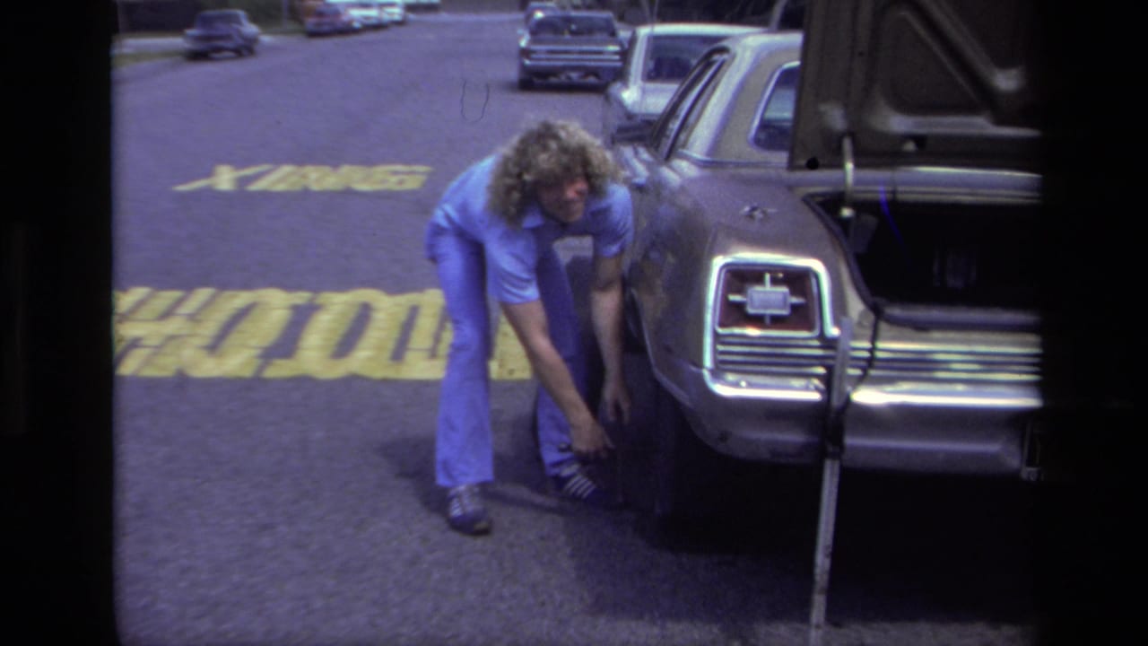 In 1979 Laguna Beach, a curly-haired man works near a car wheel with the trunk open beside a marked street.