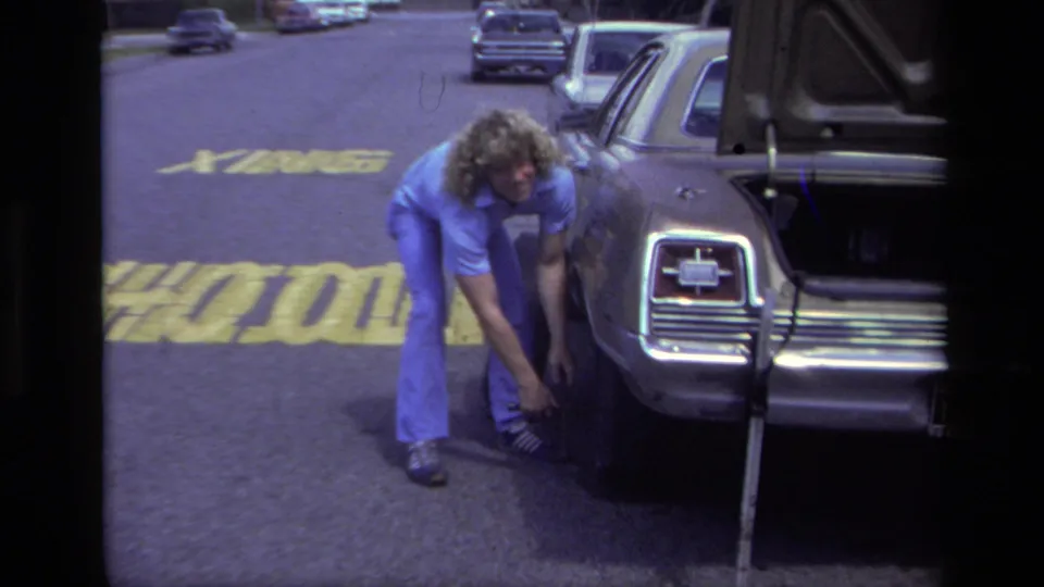 In 1979 Laguna Beach, a curly-haired man works near a car wheel with the trunk open beside a marked street.