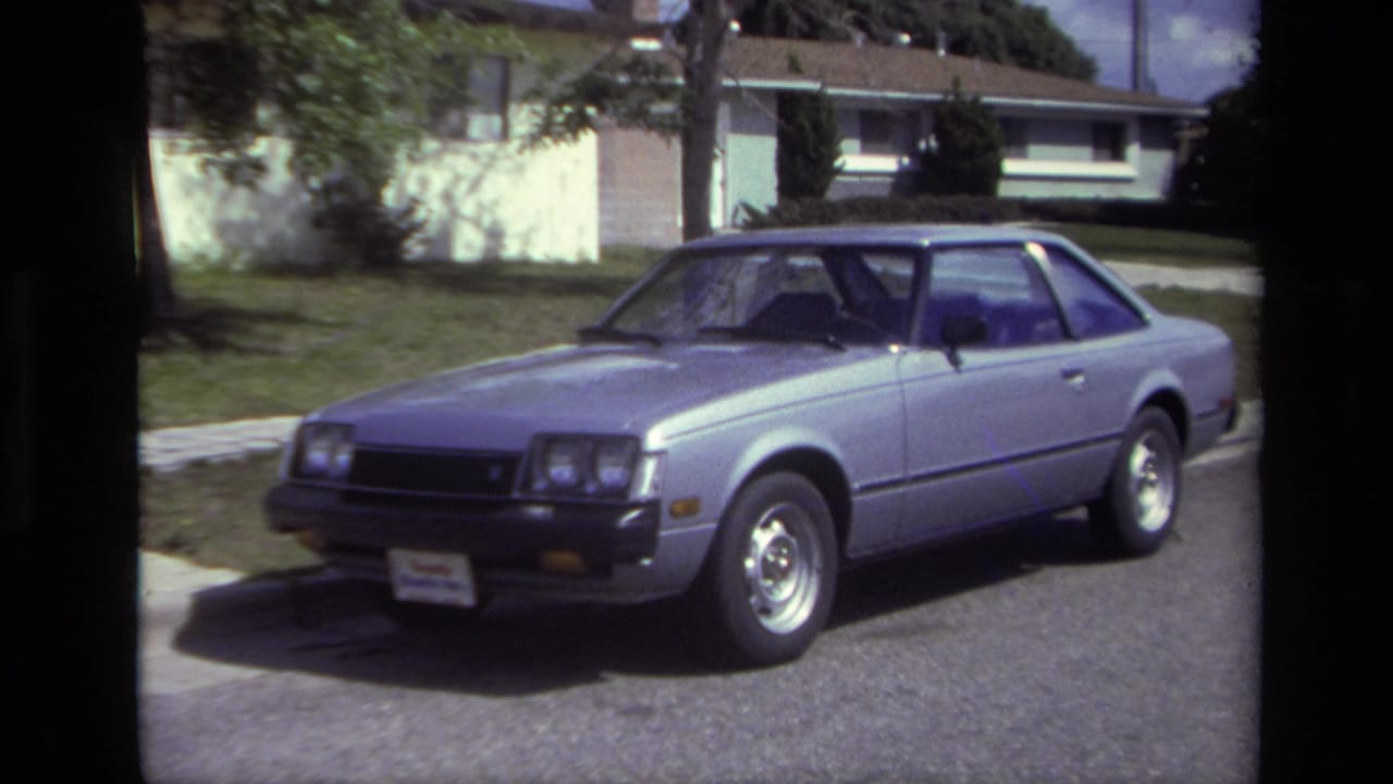 Silver two-door coupe parked at a curb in front of a lawn and low house.