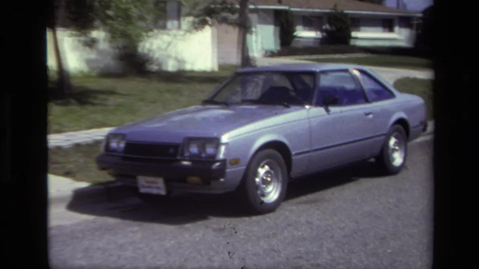 Silver 1970s coupe parked curbside in Laguna Beach, seen from the front side with pavement, lawn, trees, and a low house behind it.