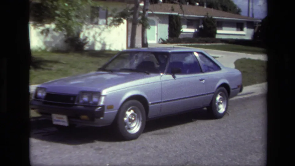 Closer Super 8 frame of the silver coupe on a residential Laguna Beach street, with front wheel, headlights, curb, and lawn visible.
