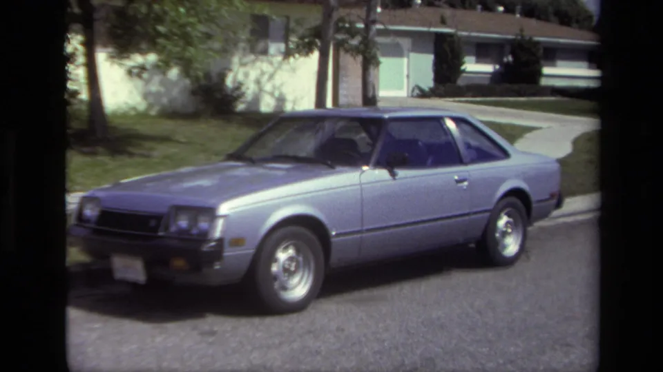 Silver 1970s coupe parked on a Laguna Beach residential street, with curb, pavement, lawn, driveway, and low house behind it.