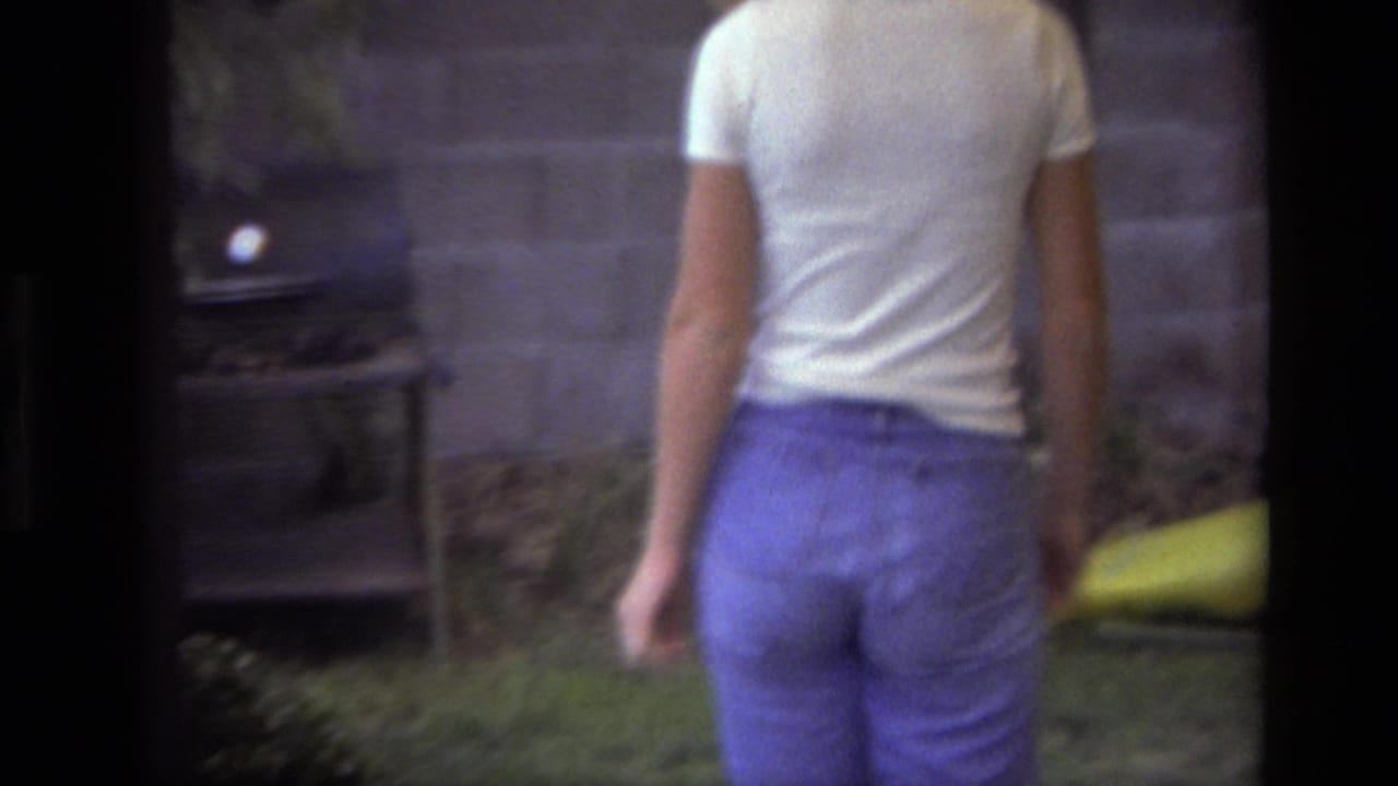 In a Laguna Beach backyard in 1979, a woman in a white t-shirt and blue pants walks away from the camera near a wall and shrubs.