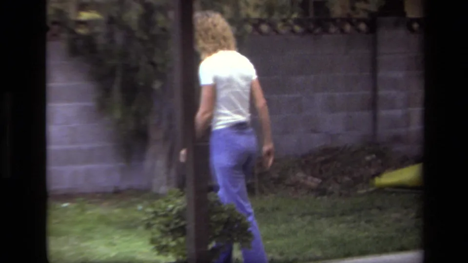 The woman continues across the Laguna Beach backyard, moving toward the far side of the lawn near shrubs and a cinder block wall.
