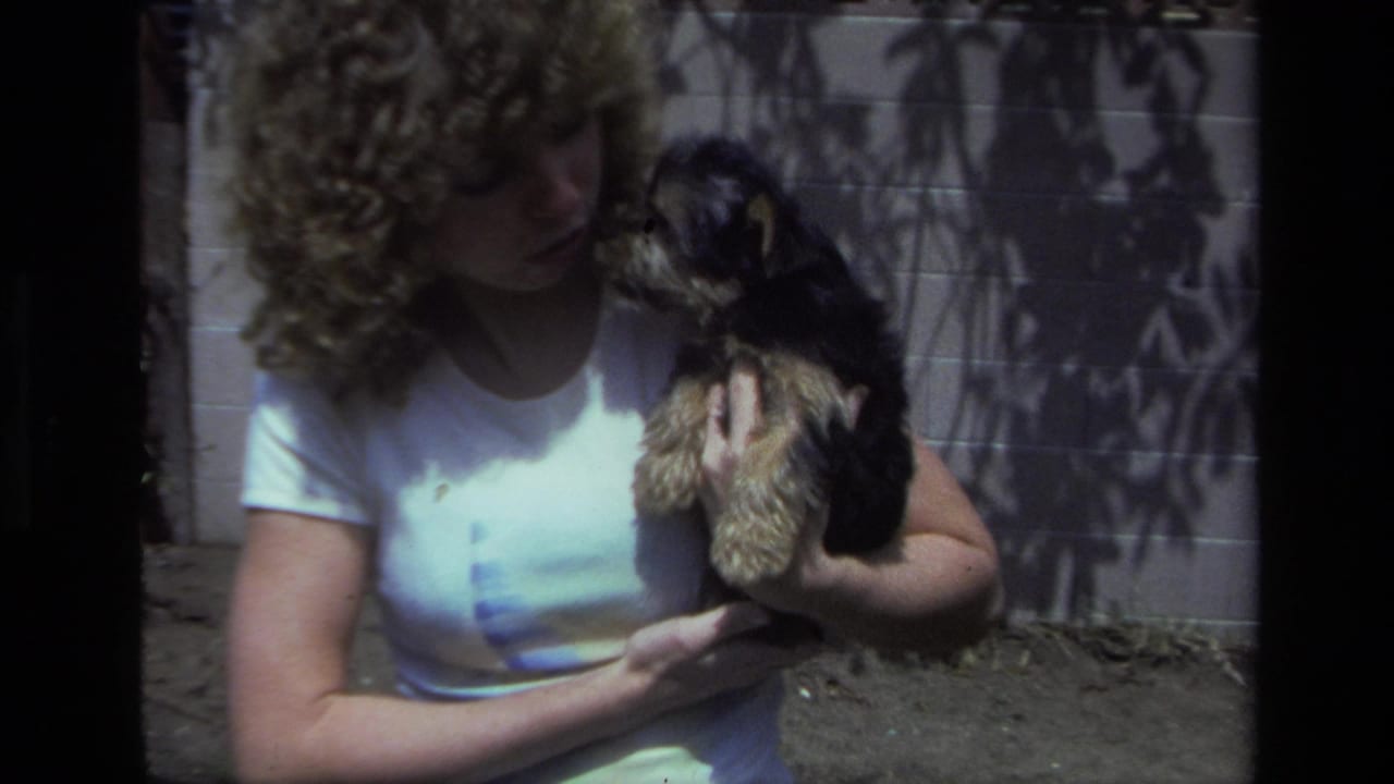 In Laguna Beach home-movie footage from 1979, a young woman cradles a small black and tan puppy near a pale wall in dappled sunlight.