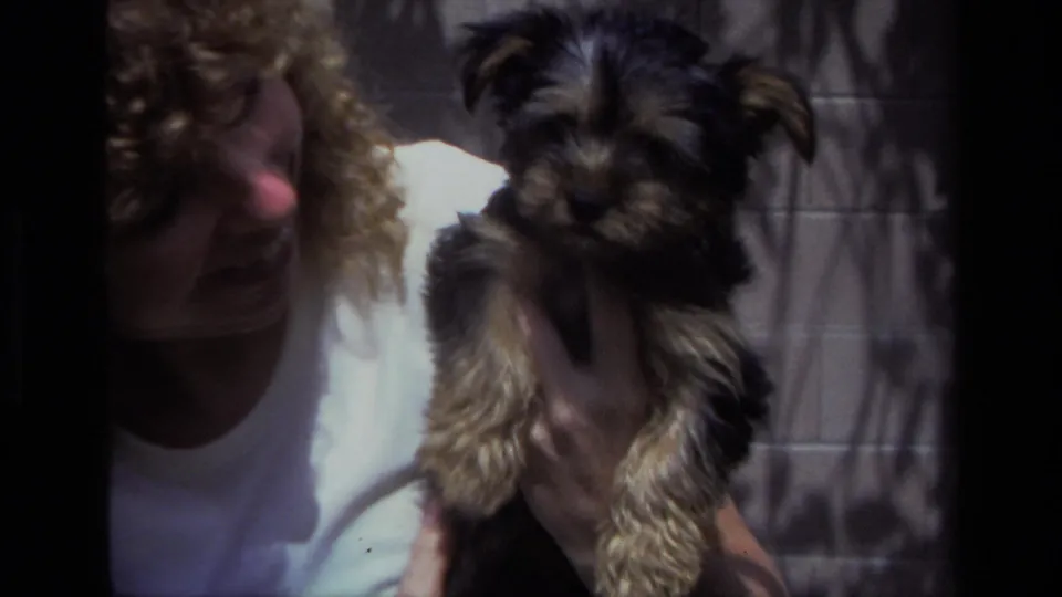 A closer 1979 8mm frame shows the black and tan puppy facing the camera while the woman holds it beside a sunlit backyard wall.