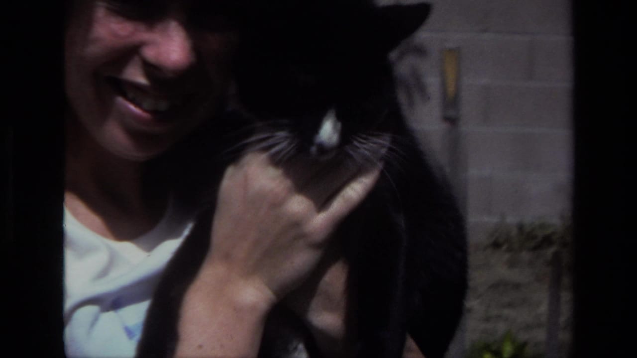 Smiling woman holds a black-and-white cat close to her face in a sunlit yard.