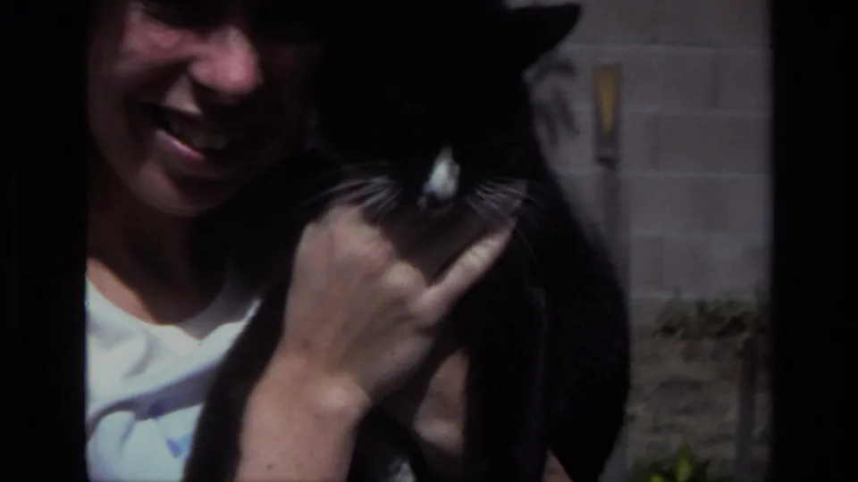 A smiling woman in 1979 Laguna Beach home-movie footage holds a black-and-white cat close beside a backyard wall.