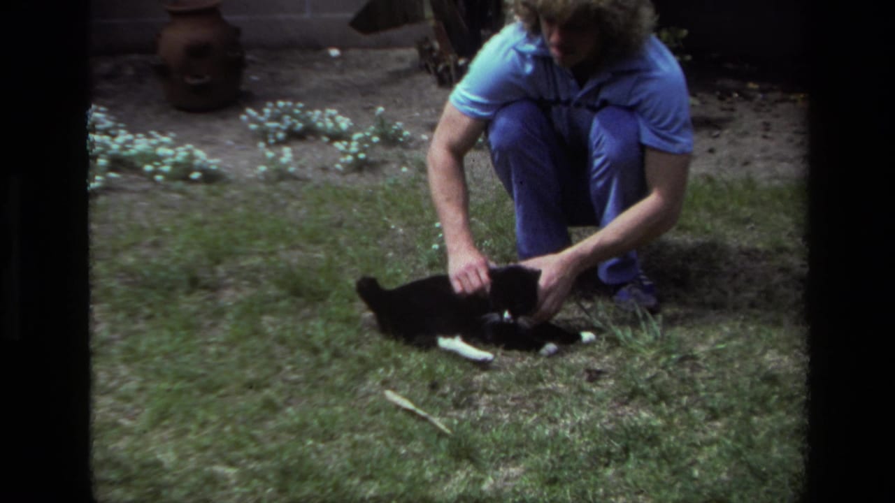 In 1979 Laguna Beach home-movie footage, a curly-haired adult in blue clothes crouches beside a black and white cat on grass.