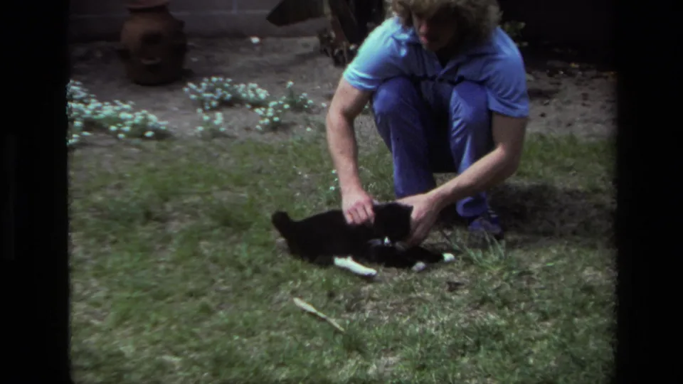 In 1979 Laguna Beach home-movie footage, a curly-haired adult in blue clothes crouches beside a black and white cat on grass.