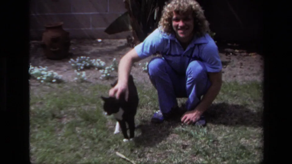 A smiling curly-haired adult crouches in a Laguna Beach backyard beside a black and white cat in faded 8mm home-movie footage.