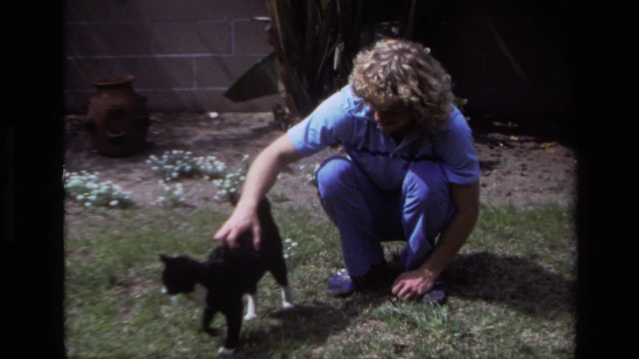 In a 1979 Laguna Beach backyard, a man in blue clothing crouches and pets a black-and-white cat on the lawn.