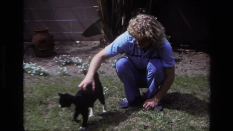 In a 1979 Laguna Beach backyard, a man in blue clothing crouches and pets a black-and-white cat on the lawn.