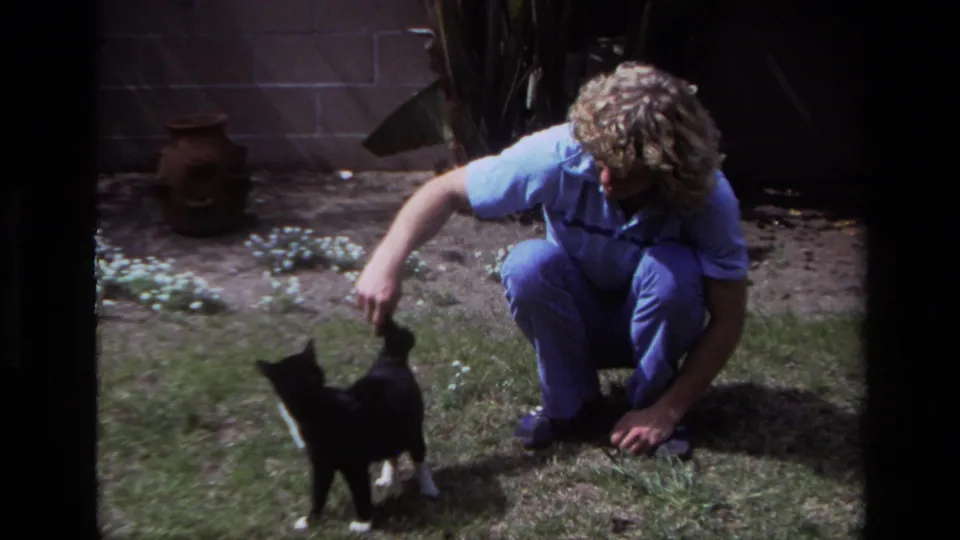 A man in blue 1970s-style casual wear pets the cat near small white flowers in the Laguna Beach backyard.