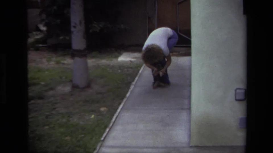 A person bends closely over a small puppy on a concrete walk in the supported 1979 Laguna Beach residential setting.