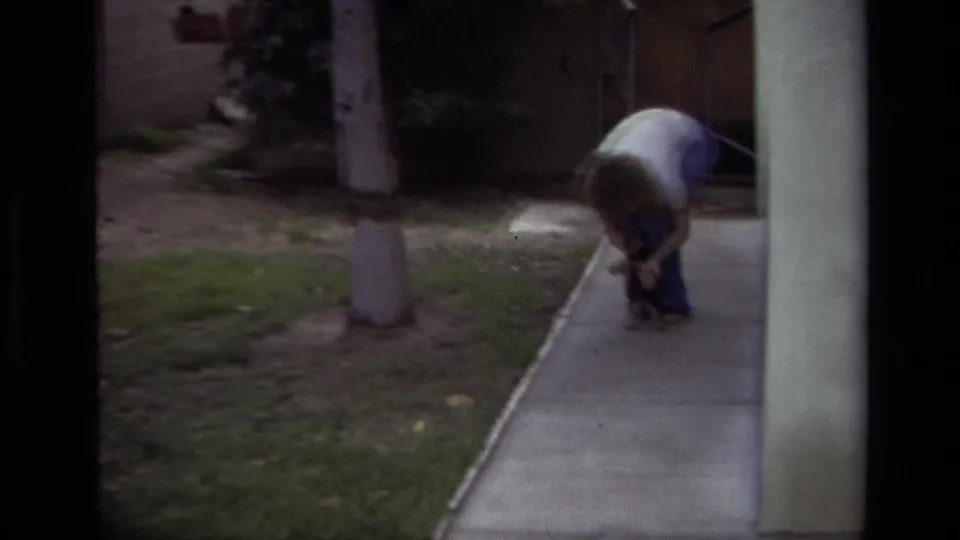 The home-movie frame shows the person still bent over the puppy along a sidewalk, with grass, a tree, and a stucco column nearby.