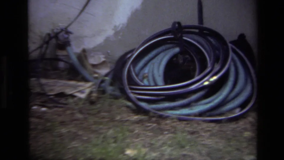1979 Laguna Beach home-movie frame with a close backyard view of a coiled hose, grass, wall, and leaves.