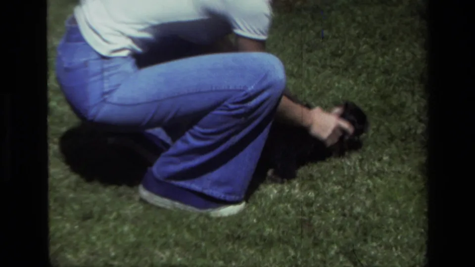 An adult wearing blue jeans and a white shirt crouches on the grass while handling a small puppy in Laguna Beach, 1979.