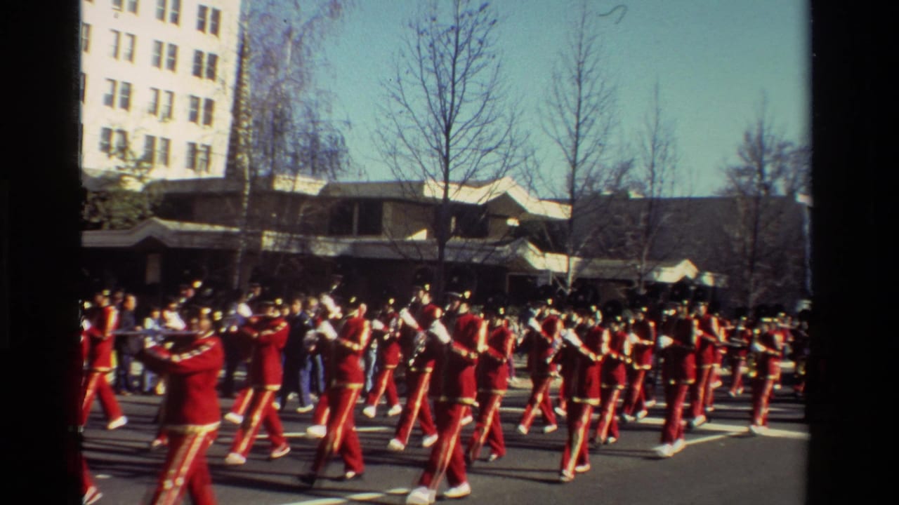 1981 California home-movie frame showing red-uniformed marching band members performing on a city street.