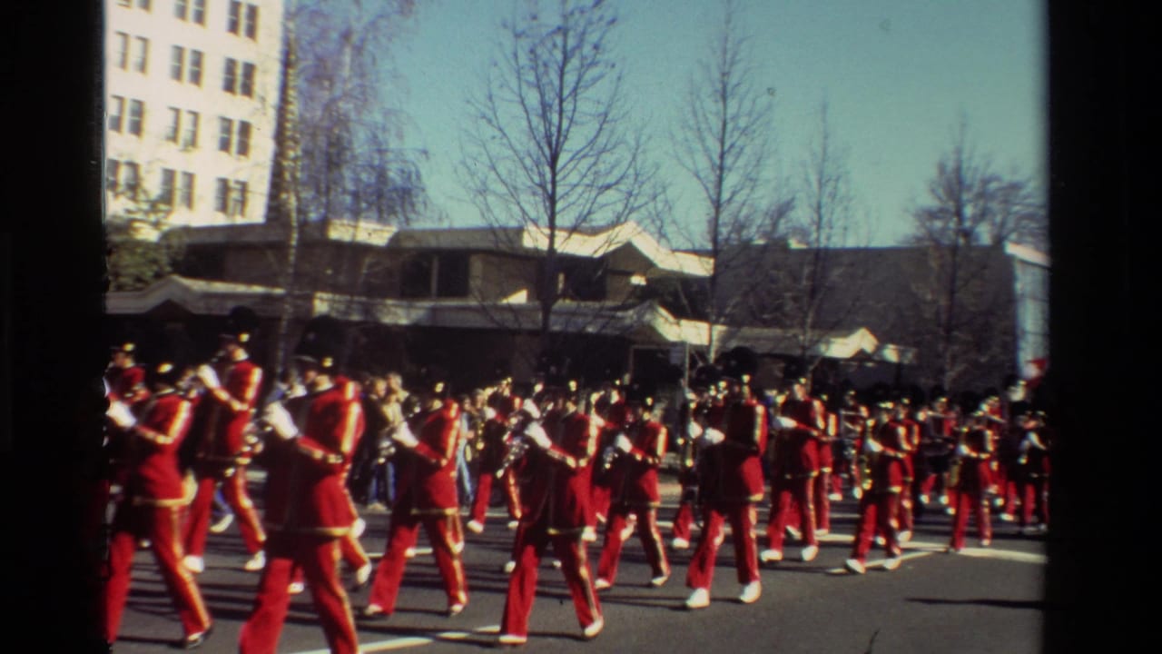 Red-uniformed musicians march in formation past spectators and buildings.