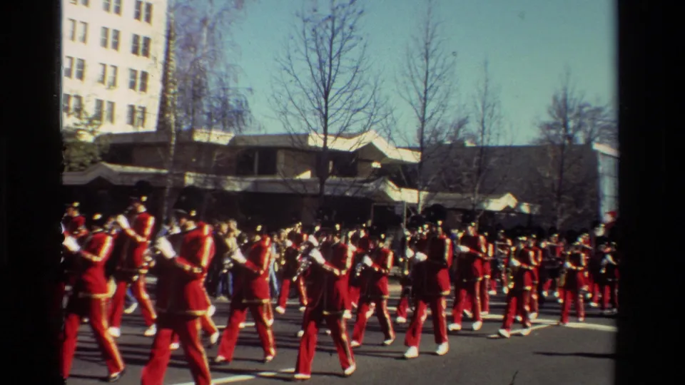 A curbside 8mm view of a California parade with musicians in red uniforms moving across the frame.