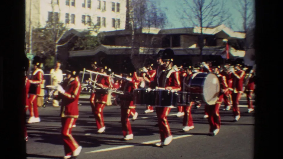 1981 California parade footage showing percussion and brass players in red marching uniforms near the curb.