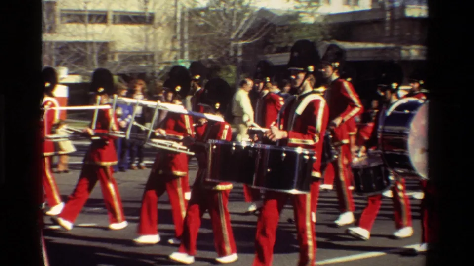 Close 8mm frame of red-uniformed marching band members with drums and brass instruments in a California street parade.