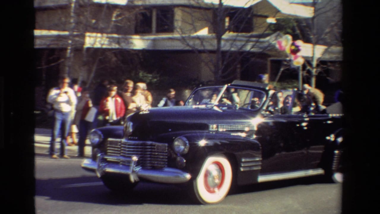 Black vintage convertible with whitewall tires rolls past spectators and balloons during the California parade sequence.