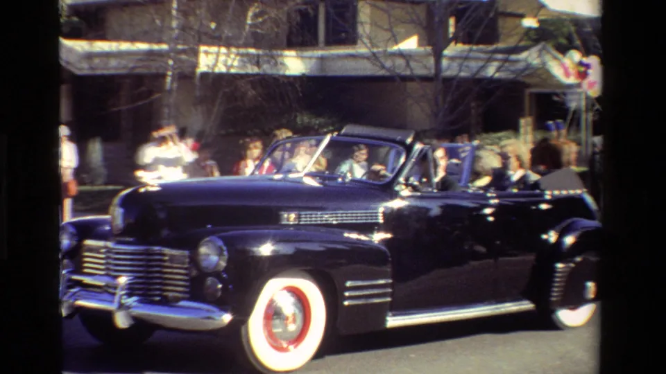 A black 1940s-style convertible with passengers passes a curbside crowd in the 1981 California parade footage.