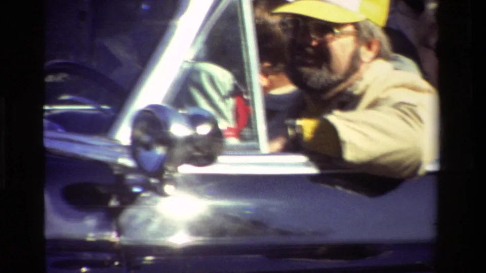 Close handheld view of a man in a yellow cap near the window of the black convertible during the parade.