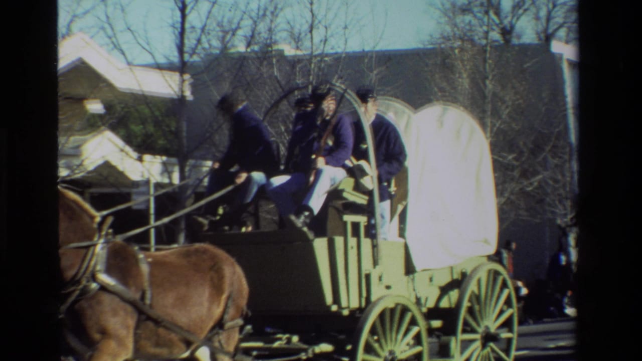 Horse-drawn covered wagon with riders in dark clothing passing along a suburban street.