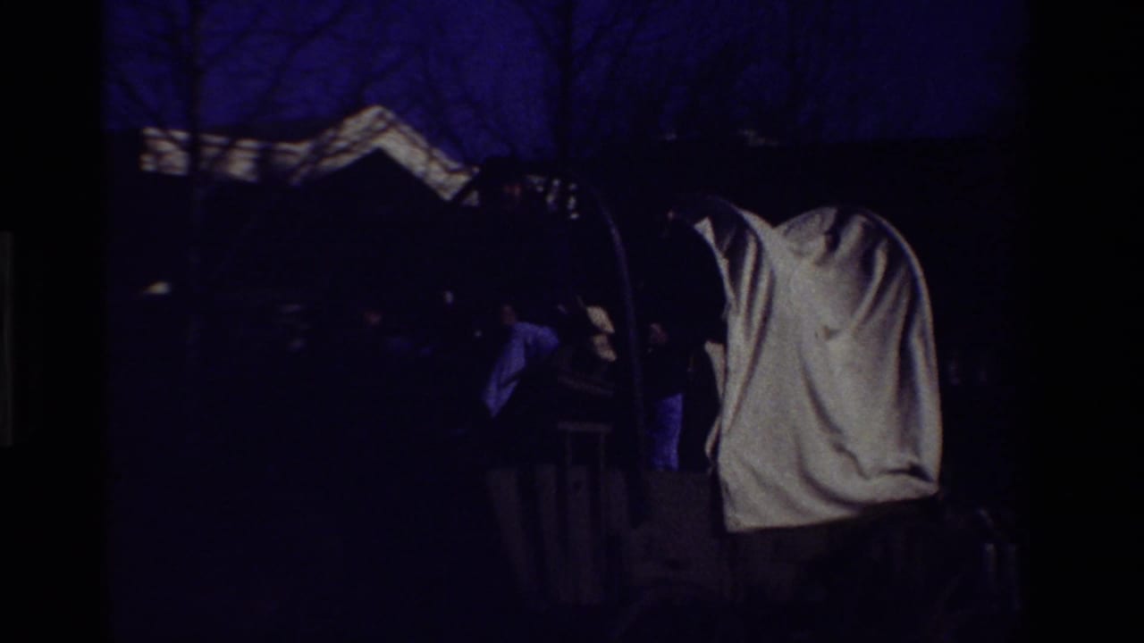 Dim frame of a covered wagon canopy and dark parade figures near trees.