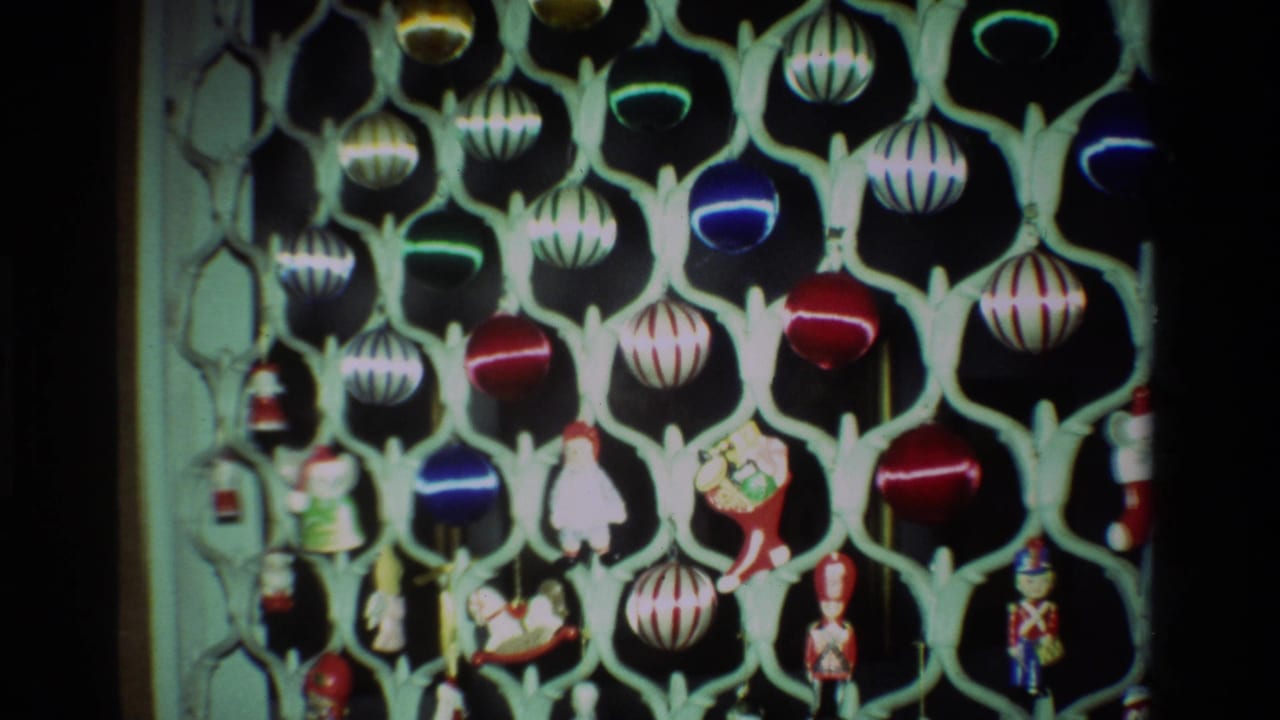 1981 California home-movie frame with a fuller view of hanging Christmas baubles, striped ornaments, and toy-like figures on a pale stand.