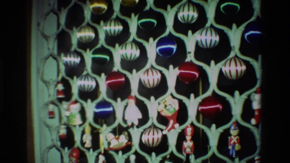 1981 California home-movie frame with a fuller view of hanging Christmas baubles, striped ornaments, and toy-like figures on a pale stand.