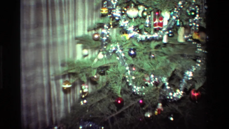 Close 8mm view of the Christmas tree branches, with tinsel, small ornaments, a red bauble, and striped curtains in the background.