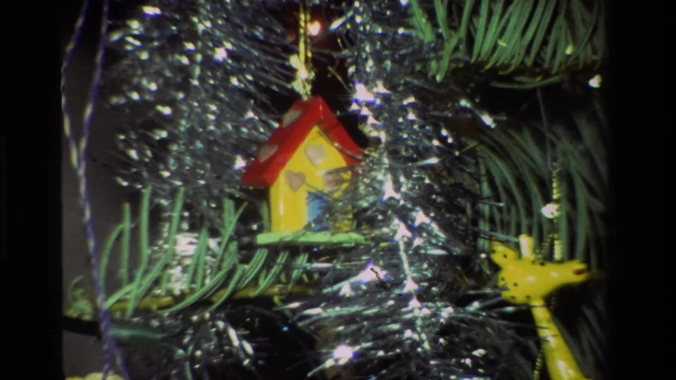 1981 California home-movie frame showing a red-roofed birdhouse ornament, yellow bird, green branches, and silver tinsel.