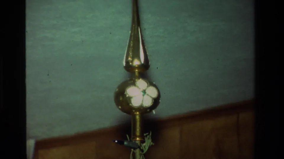 Gold Christmas tree topper with white flower decoration, seen in soft 8mm focus against an interior wall.