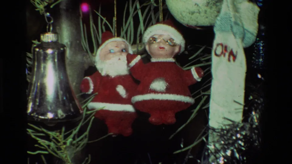 Close 1981 California 8mm view of two plush Santa ornaments hanging near a silver bell and pale stocking-like ornament on a Christmas tree.