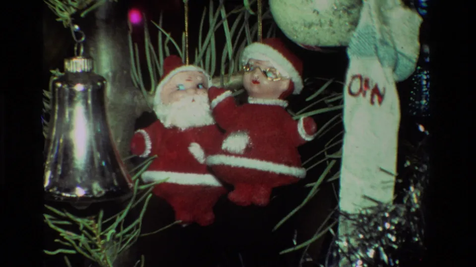 Close 1981 California 8mm frame of plush Santa figures, a silver bell, and a pale ornament hanging in dense Christmas tree branches.
