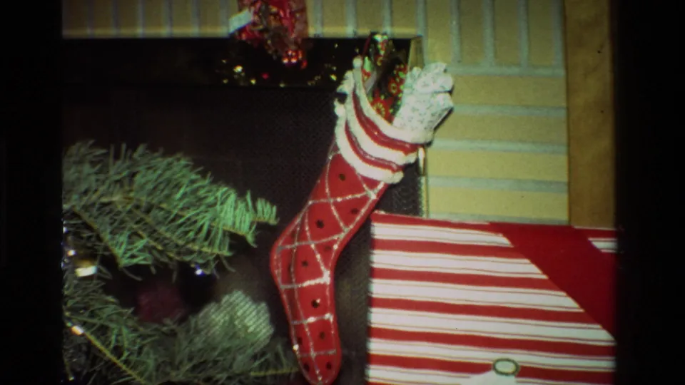 1981 California home-movie frame showing a red stocking stuffed with presents beside a fireplace and striped gift box.