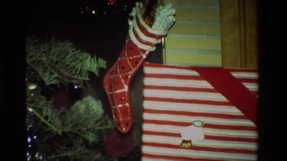 1981 California home-movie frame showing a red Christmas stocking, tree branches, ornaments, and a red-and-white striped present.