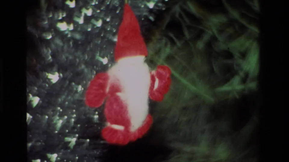 Close 1981 California home-movie frame of a small red-and-white Santa-like ornament hanging among dark green Christmas tree branches.