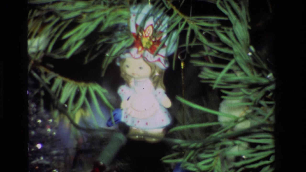 Small doll ornament in a floral hat hanging among Christmas tree branches.