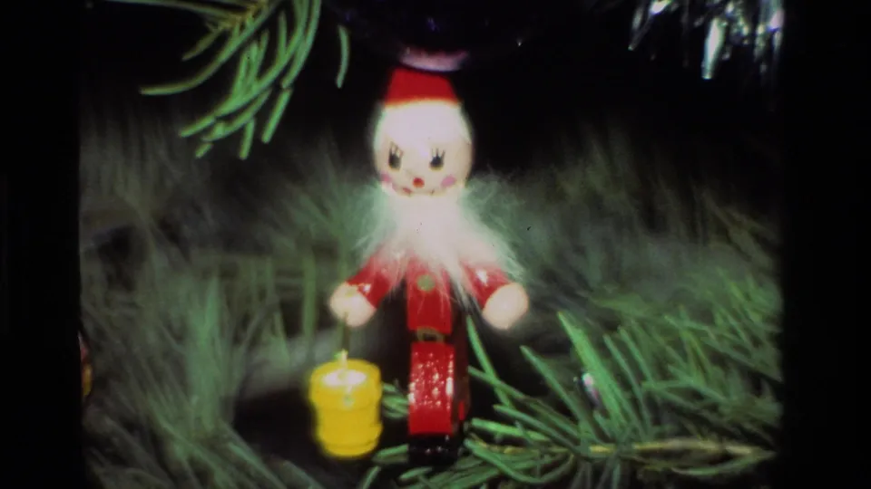 Close view of a red elf ornament with a white beard and yellow lantern hanging on a Christmas tree in 1981 home-movie footage.