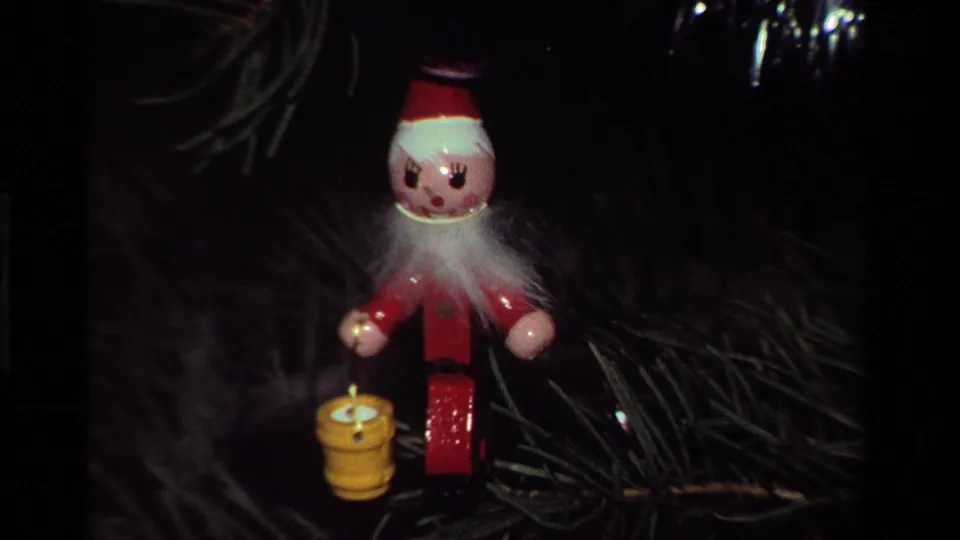 A red elf ornament holding a yellow lantern appears against darker pine branches, with 8mm grain and soft holiday lighting.