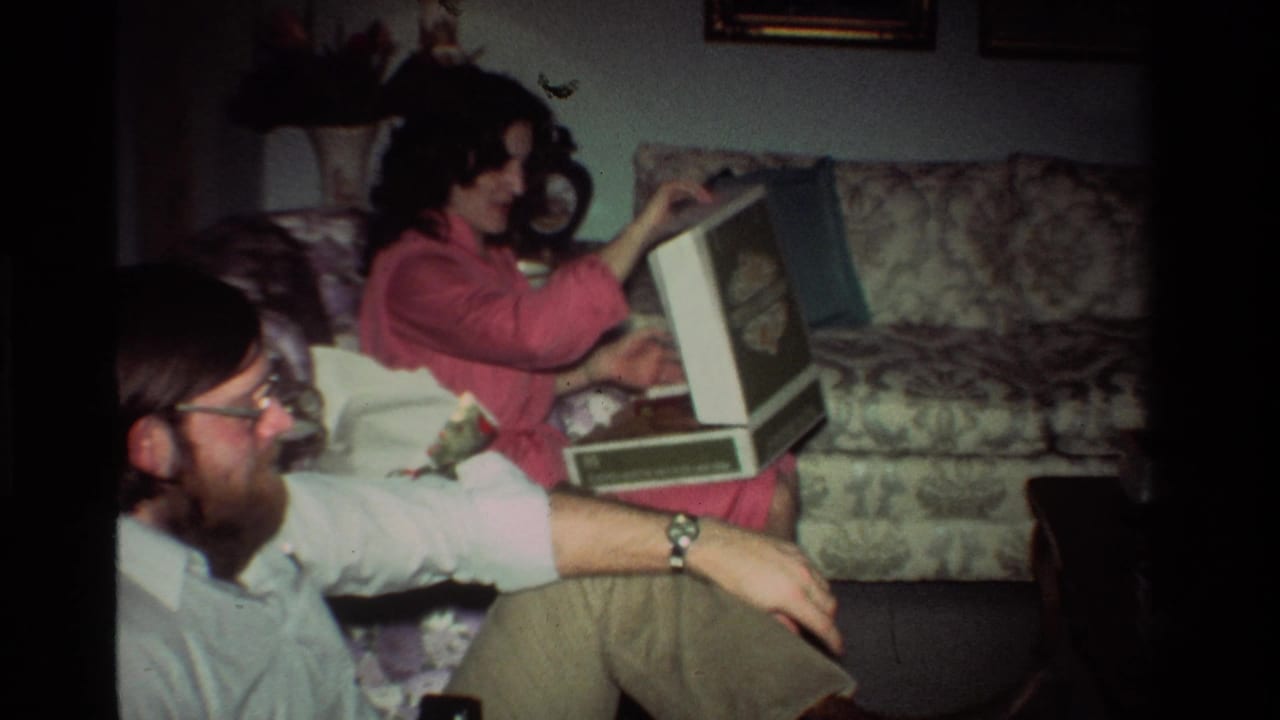 Woman in pink opens a gift box on a patterned couch while a man with glasses sits nearby.