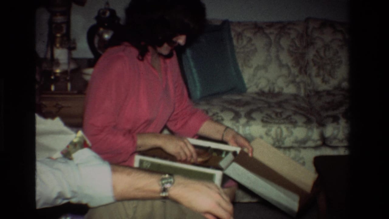 The woman in pink leans over an open gift box in the California home interior, with the seated man’s arm and profile partly visible nearby.
