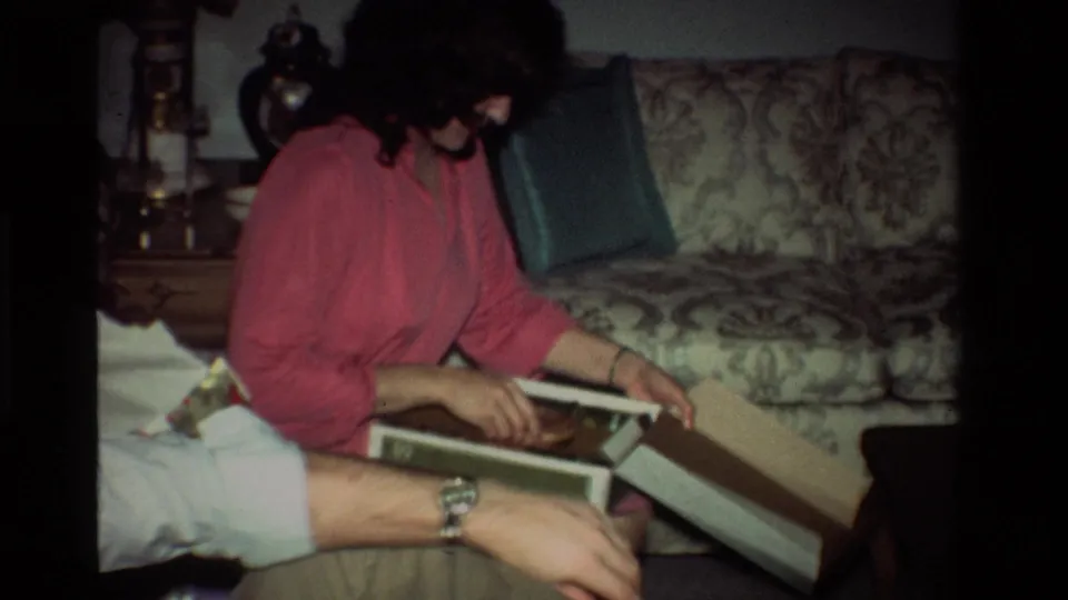 The woman in pink leans over an open gift box in the California home interior, with the seated man’s arm and profile partly visible nearby.